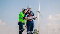 Mann und junge Frau in Arbeitskleidung mit Helm gucken vor Windrad in einen Plan