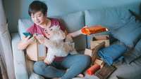 Asiatische Frau sitzt mit Hund auf dem Sofa und öffnet zufrieden viele Pakete