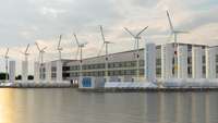 Windräder, Solarpanele und Wasserstofftanks vor einem Industriegebäude an einem Gewässer