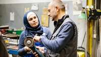 Ein Techniker erklärt einer weiblichen, muslimischen Auszubildenden in einer Werkstatt eine Schleifmaschine
