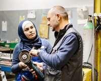 Ein Techniker erklärt einer weiblichen, muslimischen Auszubildenden in einer Werkstatt eine Schleifmaschine