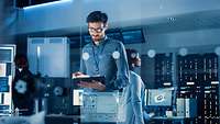 Ein Mitarbeiter mit Brille arbeitet an einem Tablet, im Hintergrund sind weitere Bildschirme und Mitarbeitende zu erkennen