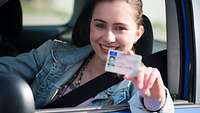 Junge Dame sitzt im Auto und hält ihren Führerschein in die Kamera