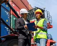 Mann im Anzug und Frau mit Warnweste, beide mit Helm, stehen mit einem Klemmbrett vor Containern 