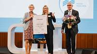 Ein Mann und eine Frau überreichen einer weiteren Frau auf der Bühne eine Urkunde und einen Blumenstrauß 