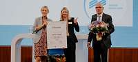 Ein Mann und eine Frau überreichen einer weiteren Frau auf der Bühne eine Urkunde und einen Blumenstrauß 