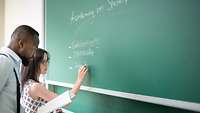 Zwei Personen stehen an einer Tafel, eine Frau schreibt mit Kreide