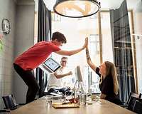 Junge Menschen an einem Schreibtisch, einer steht auf dem Stuhl und gibt seiner Kollegin "high five"