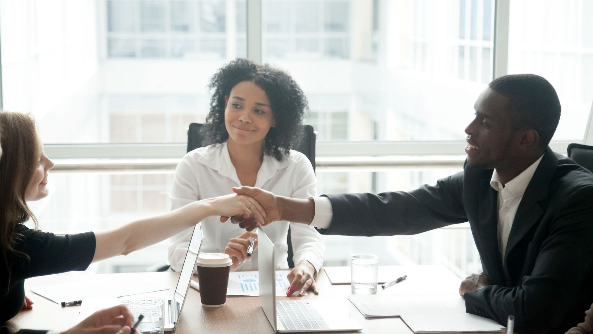 Ein schwarzer Mann und eine weiße Frauen schütteln sich quer über einen Bürotisch die Hände