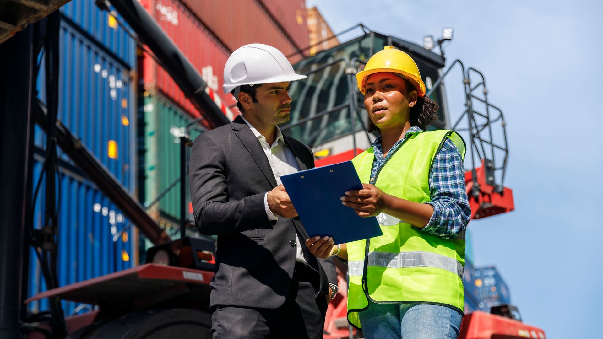 Mann im Anzug und Frau mit Warnweste, beide mit Helm, stehen mit einem Klemmbrett vor Containern 
