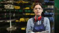 Arbeiterin mit Ohrenschützern und Handschuhen steht im Lager eines Metallbearbeitungswerkes 