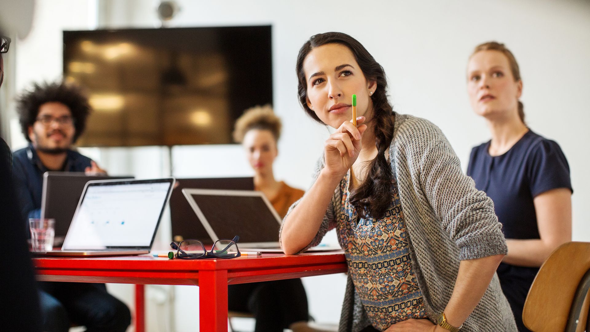 Junge Menschen sitzen an einem Tisch mit Laptops, im Vordergrund eine dunkelhaarige Frau mit Stift in der Hand