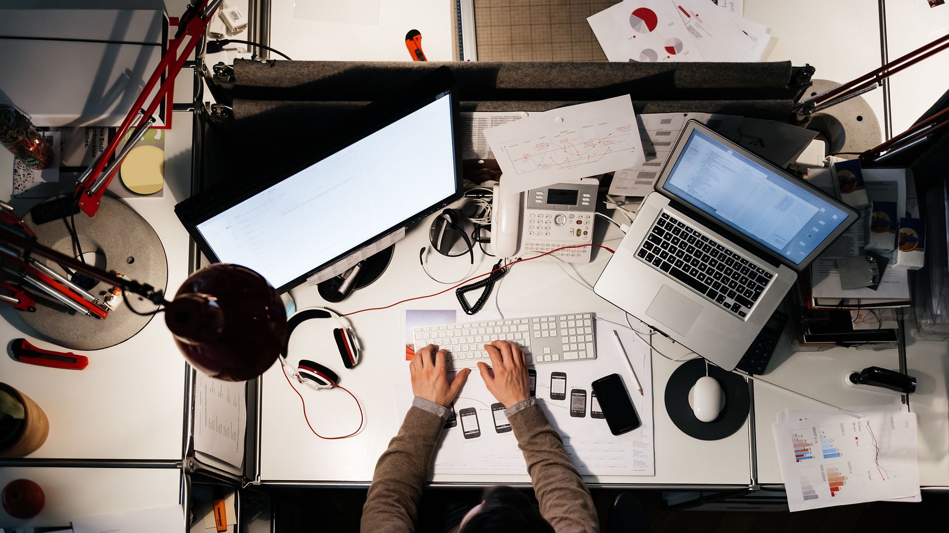 Mannn von oben an einem Schreibtisch, der mit Computern, Telefonen, Handys und weiteren Elektrogeräten bedeckt ist