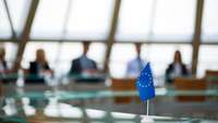 Tisch mit EU-Flagge, unscharf im Hintergrund Menschen