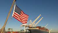 Frachter liegt an einem Hafenkai mit Kränen; im Vordergrund eine US-Flagge