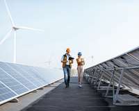 Ein Ingenieur und eine Ingenieurin wandern durch ein Feld von Solarpanelen, im Hintergrund Windräder