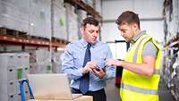 Lagerarbeiter und Manager stehen in einem Lager vor einem Karton, auf dem ein Laptop steht, und bedienen ein mobiles Datengerät