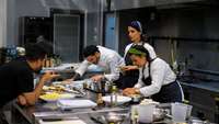 Ein Ausbilder gibt drei jungen Koch-Azubis, die an einem Profi-Herd ein Essen zubereiten, Tipps