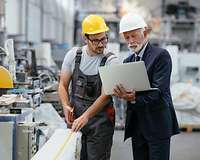 Ein Manager mit Laptop und ein Arbeiter stehen in einer Fabrik an einer Maschine