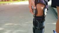 Mann mit kurzen Hosen geht eine Straße entlang, das Bein ist mit einer Kniespange bandagiert