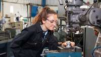 junge Frau mit Schutzbrille und Blaumann an metallverarbeitender Maschine