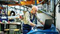 Flugzeugmechaniker steht in einer Werkhalle über einen Laptop gebeugt