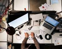 Mannn von oben an einem Schreibtisch, der mit Computern, Telefonen, Handys und weiteren Elektrogeräten bedeckt ist
