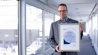 Markus Mettler Mann steht im Flur eines Unternehmens und hält eine gerahmte Urkunde in die Kamera