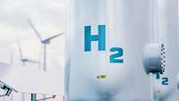 Großer Wasserstofftank mit der Aufschrift "H2", im Hintergrund unscharf Windräder und Solarpanels
