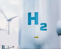 Großer Wasserstofftank mit der Aufschrift "H2", im Hintergrund unscharf Windräder und Solarpanels
