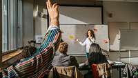 Personen sitzen in einem Klassenraum und eine Frau steht an der Tafel