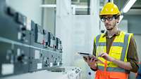 Ingenieur mit Helm, Warnweste und Tablet steht in einem Kontrollraum und checkt Solarstrom-Konverter